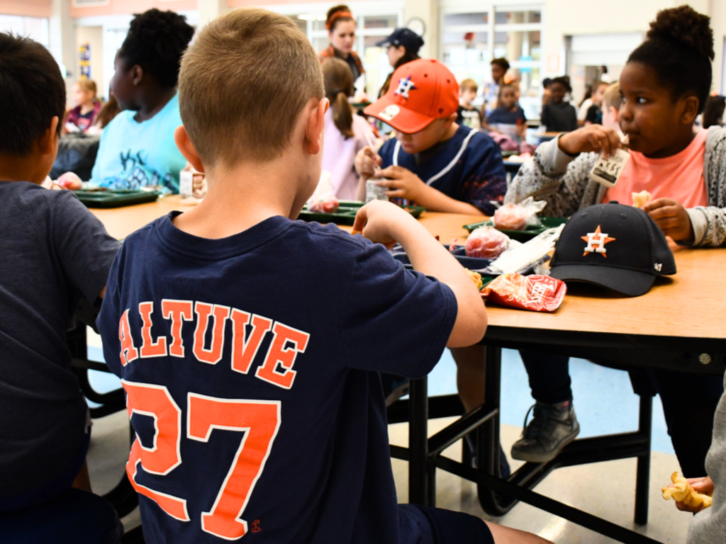 A Mitchell Elementary School student wears a Jose Altuve No. 27 shirt with a Houston Astros hat sitting next to his lunch tray, while a student across from him wears Houston Astros gear also to celebrate the Astros' World Series win.